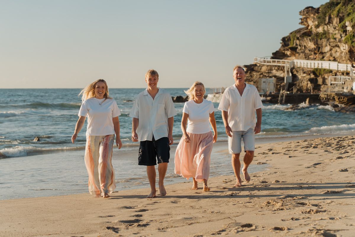 Four adults walking barefoot on a sunny beach near rocky cliffs, wearing casual white outfits on a clear day Bronte Photoshoot Sydney