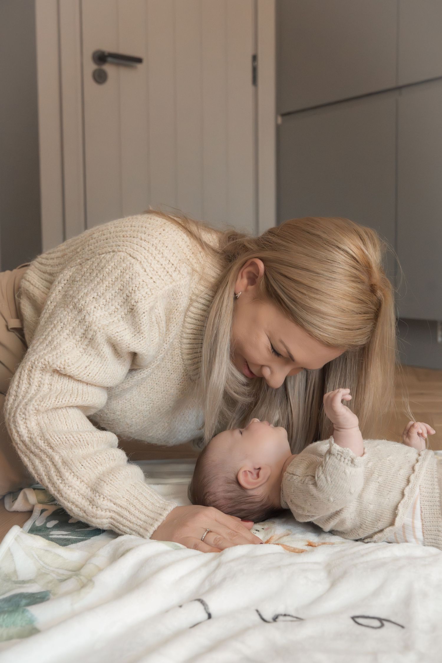 A blonde mother in a cream sweater tenderly leans over her smiling newborn baby lying on a soft blanket.