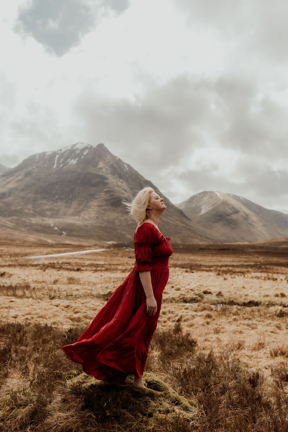 Woman in flowing red dress stands on highland moor with snow-capped mountains backdrop on cloudy day.