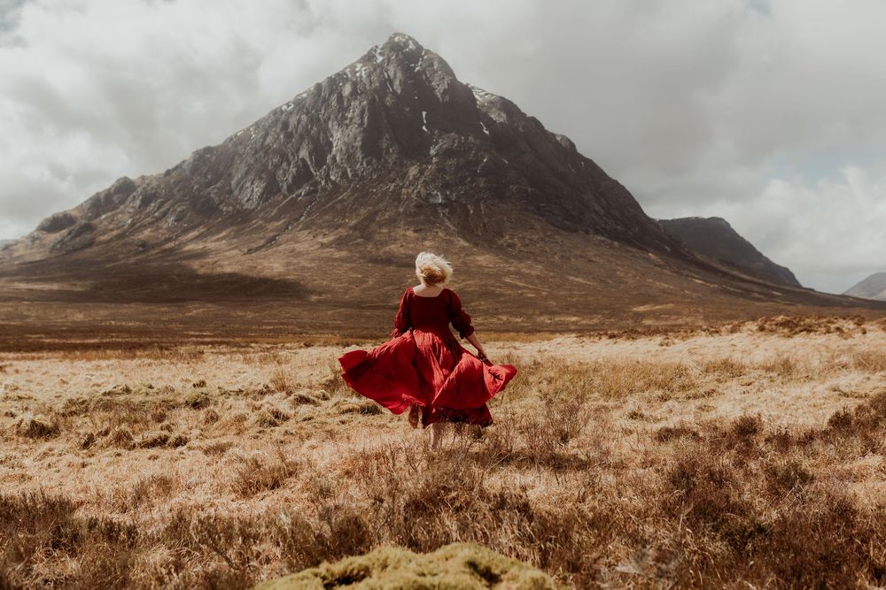 Woman in red dress twirling in Scottish Highland moorland with dramatic mountain backdrop under cloudy sky.