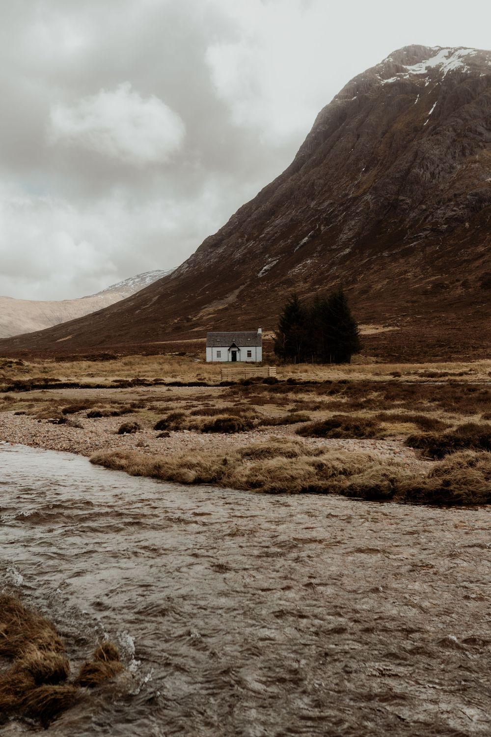 Lone white cottage beside trees in a vast Scottish Highland valley with a rocky river and snow-capped mountain under cloudy skies.