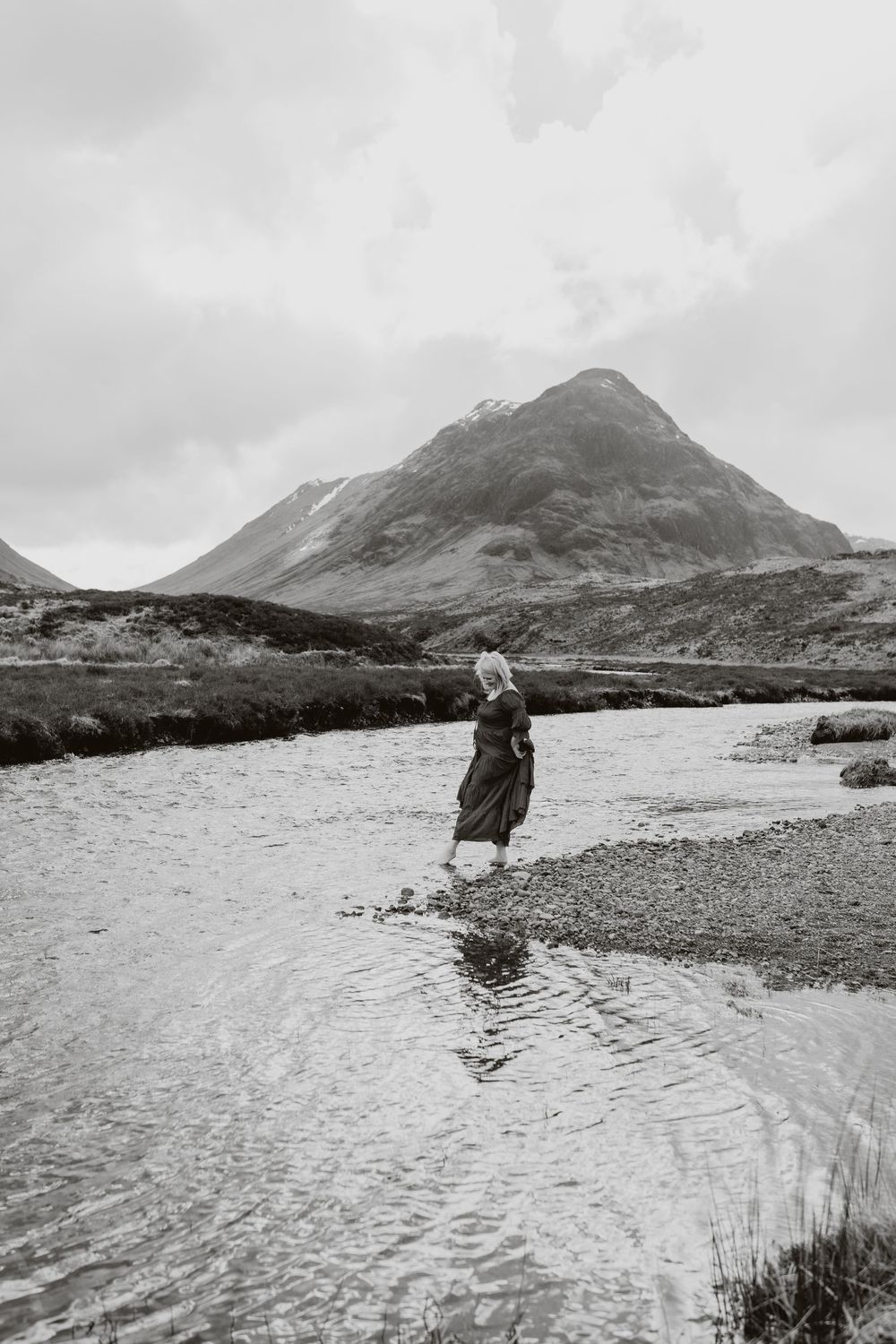 Woman in long dress wading through a shallow river with a dramatic mountain backdrop in black and white.