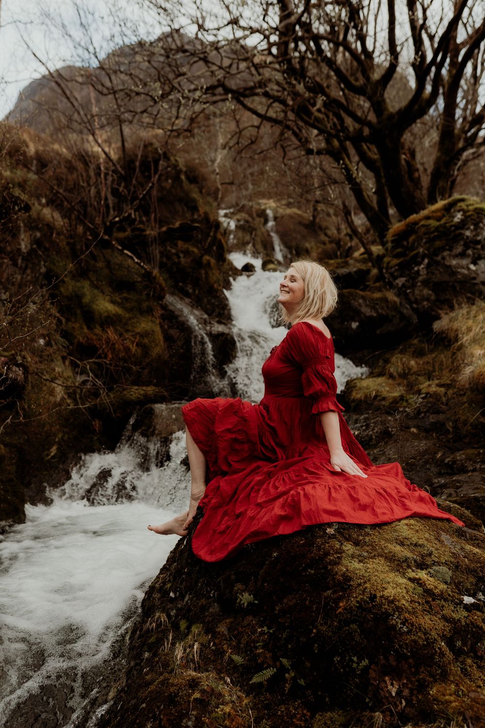 A blonde woman in a flowing red dress sits barefoot on mossy rocks beside a cascading waterfall in a rugged landscape.
