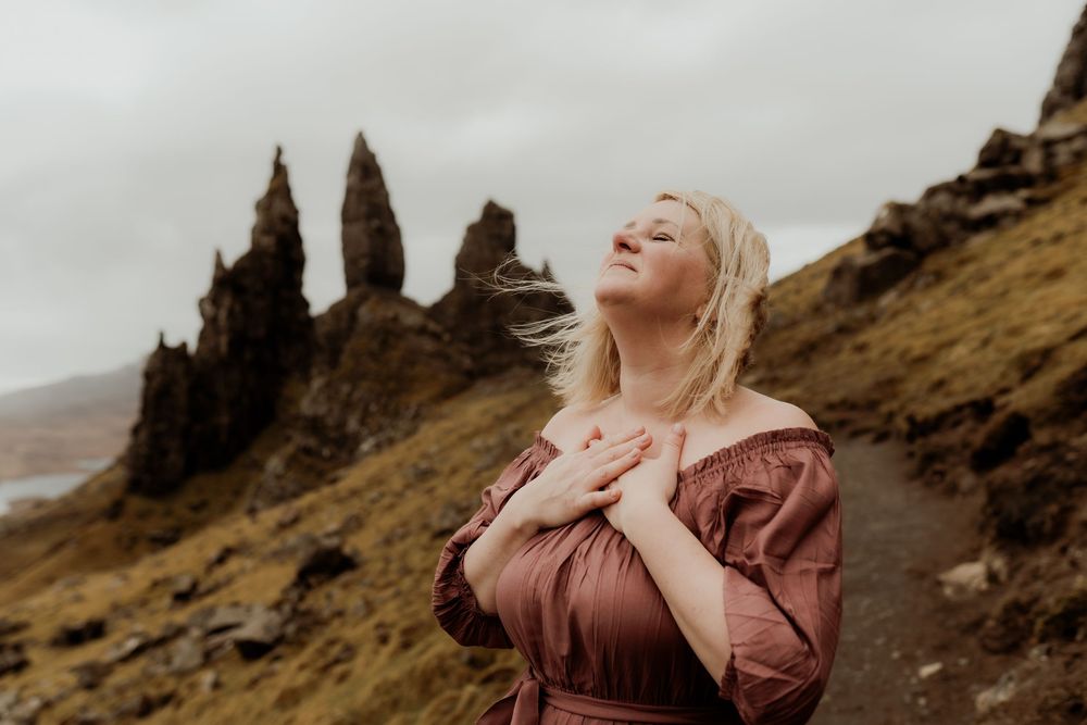 Blonde woman in dusty rose off-shoulder dress stands at Old Man of Storr, Isle of Skye, hands on chest, eyes closed.