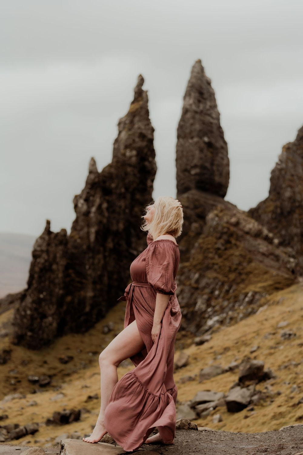 A blonde woman in a flowing dusty rose maxi dress poses barefoot among dramatic rocky spires on the Isle of Skye.