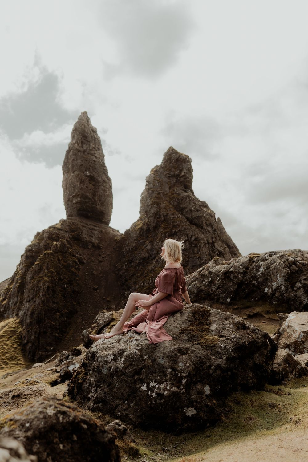 Woman in dusty rose dress sitting on rugged rocks with dramatic stone spires rising behind her under a cloudy sky.