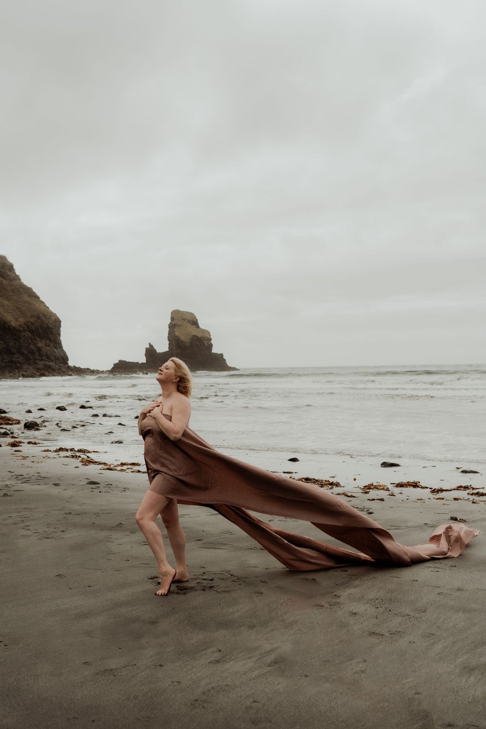 Woman wrapped in flowing brown fabric poses on a misty beach with rocky sea stacks in the background.