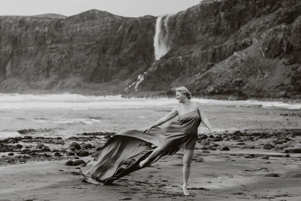 Woman in flowing dress dancing on rocky beach with dramatic waterfall and cliffs in black and white photo.