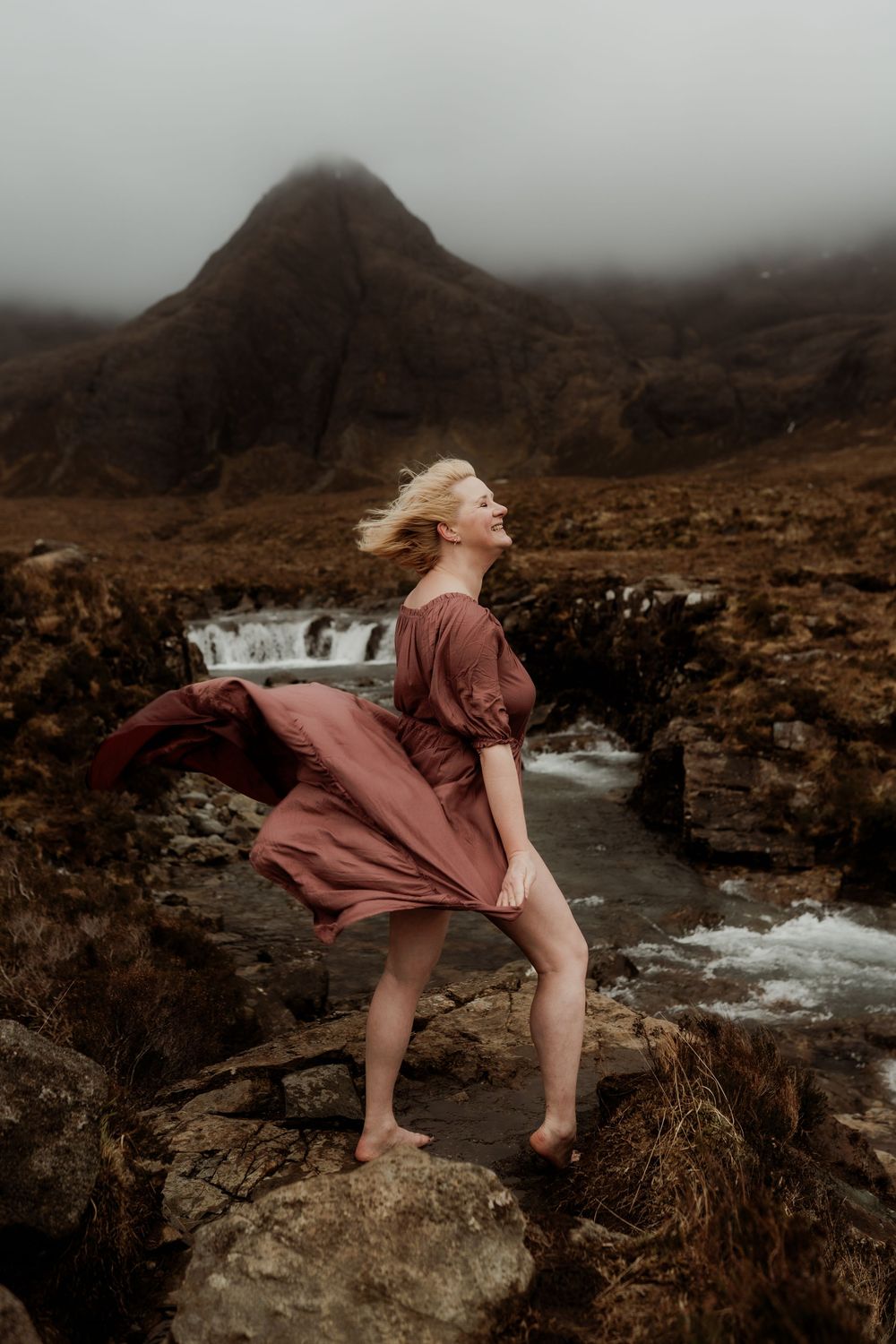 Barefoot blonde woman in flowing rust dress stands on rocks by waterfall with misty mountains backdrop.