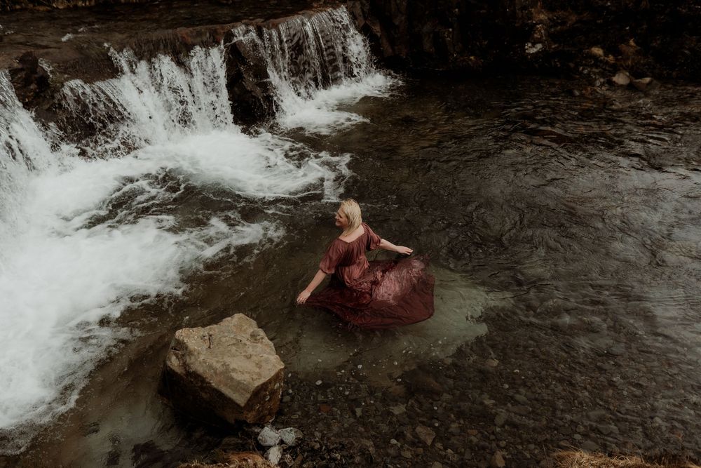 Woman in a flowing burgundy gown sitting in a rocky river stream with a cascading waterfall in the background.