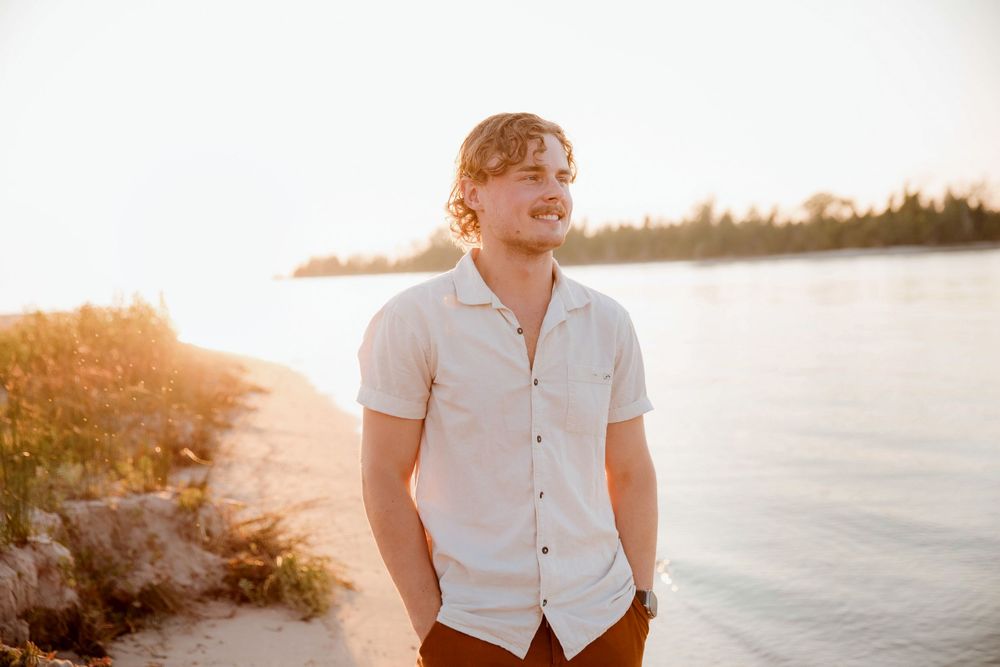 Young man in white shirt smiling on sandy beach at golden sunset with calm water and trees in background.