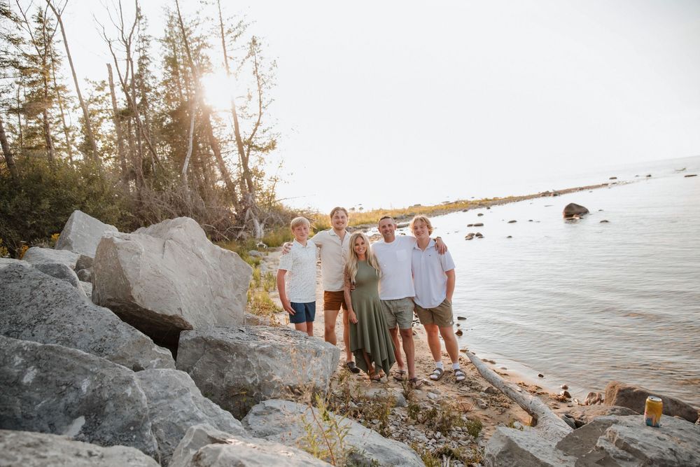 Family of five posing on rocky shoreline with sunlit trees and calm water in background at Fisherman's Island State Park in Charlevoix.