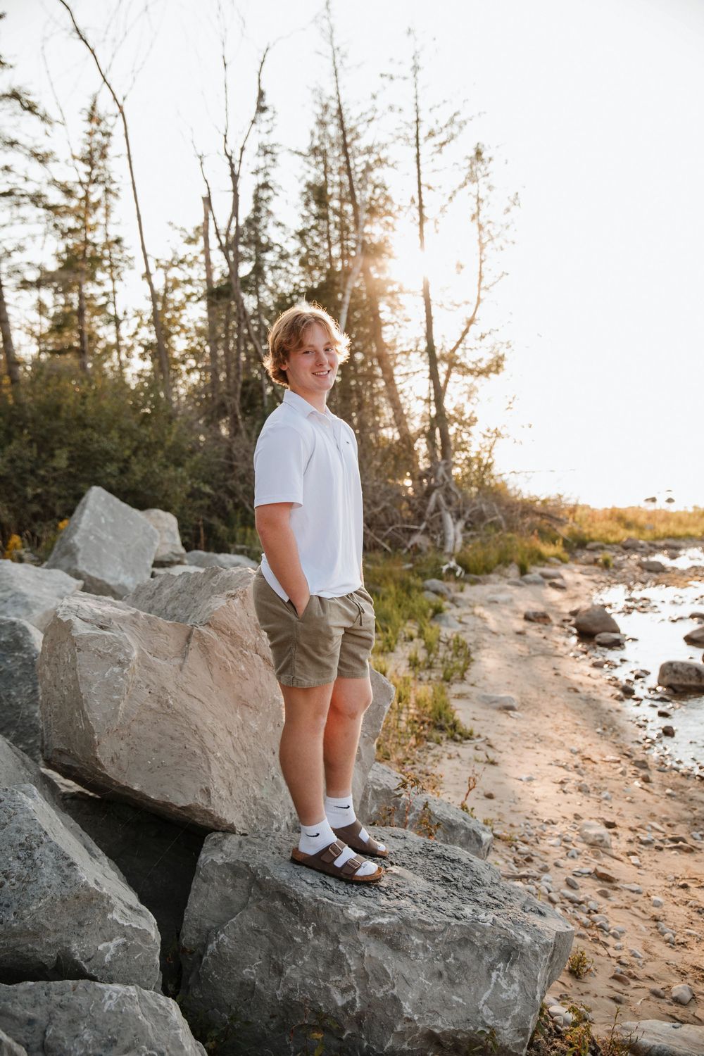 Teen boy in white polo and khaki shorts standing on rocks by a sandy shoreline at golden hour sunset.
