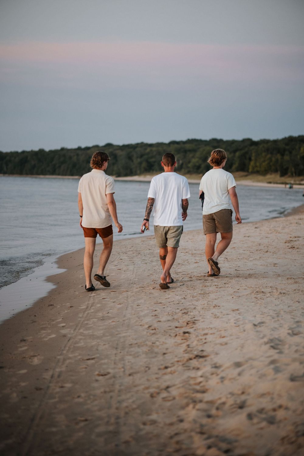 Three men walking along a sandy beach at sunset wearing white shirts and shorts.