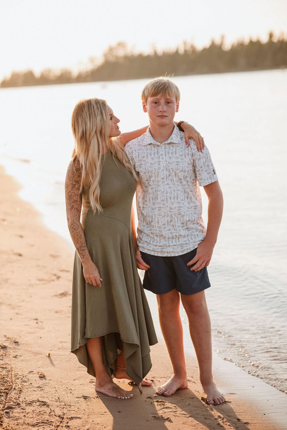 A woman in a green dress and a teenage boy pose together on a sandy beach at golden hour.