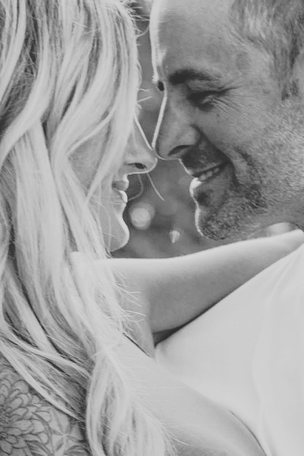 Black and white photo of a smiling couple facing each other closely, woman with blonde hair and tattoos, romantic moment.