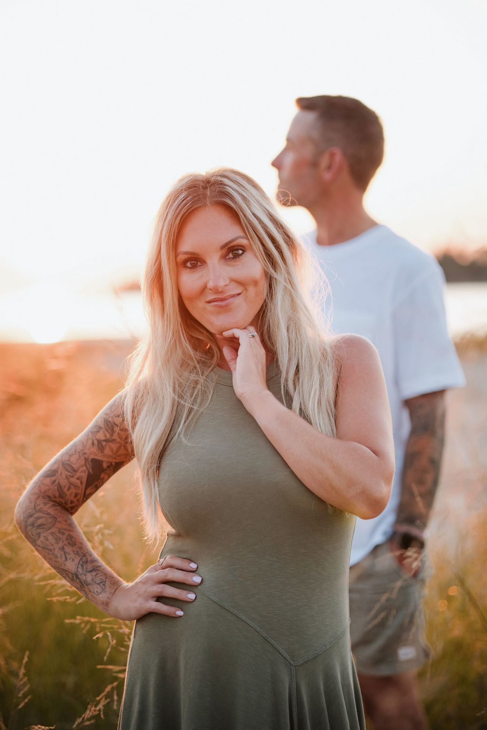 Tattooed blonde woman in green dress poses at golden hour with a man in white t-shirt in a field.