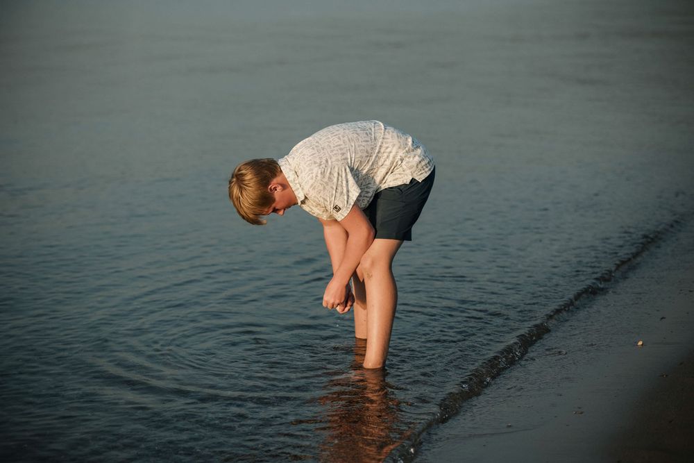 Young boy wading in shallow ocean water at the beach during sunset looking for rocks