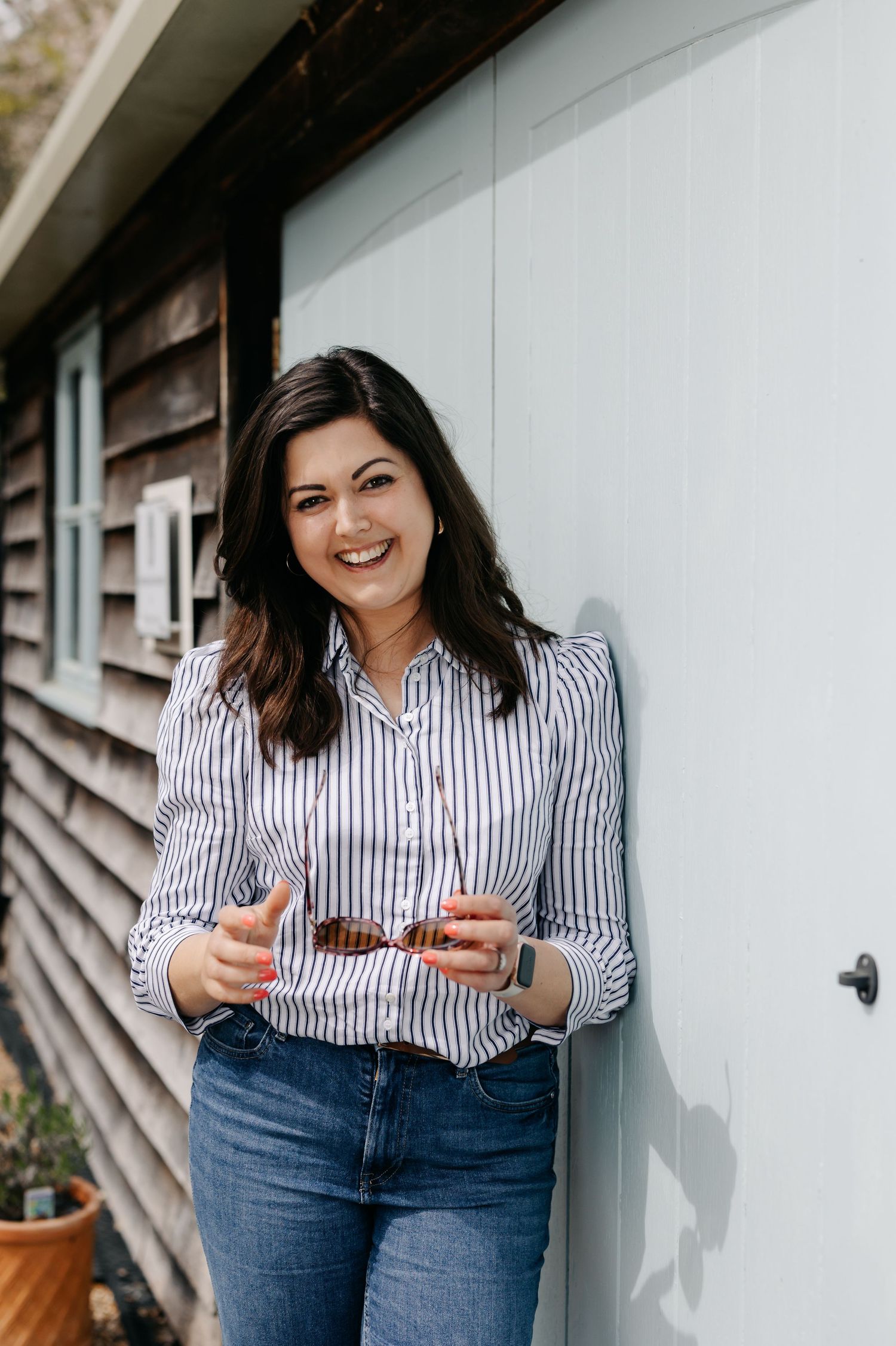 Smiling brunette woman in striped shirt and jeans leaning against a light blue wall outdoors.