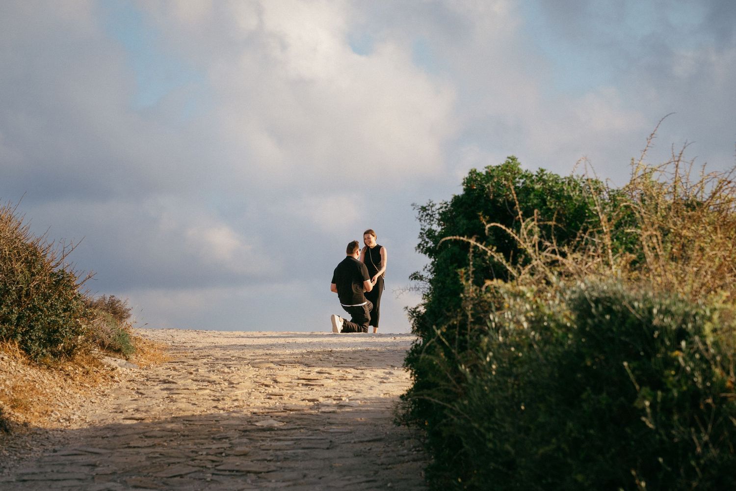 Couple walking along a sandy beach path surrounded by green shrubs under a cloudy sky, getting photographed during secret engagement.