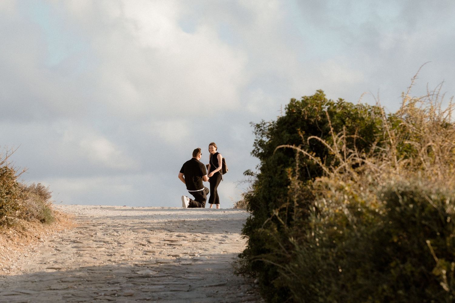 Couple walking along a sandy beach path surrounded by green shrubs under a cloudy sky, getting photographed during secret engagement.
