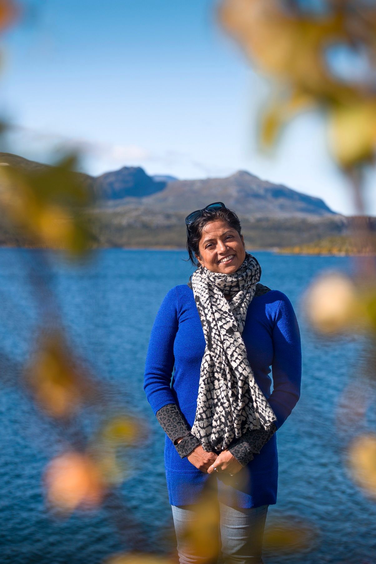 Woman in blue coat and scarf standing by a mountain lake on a sunny autumn day.