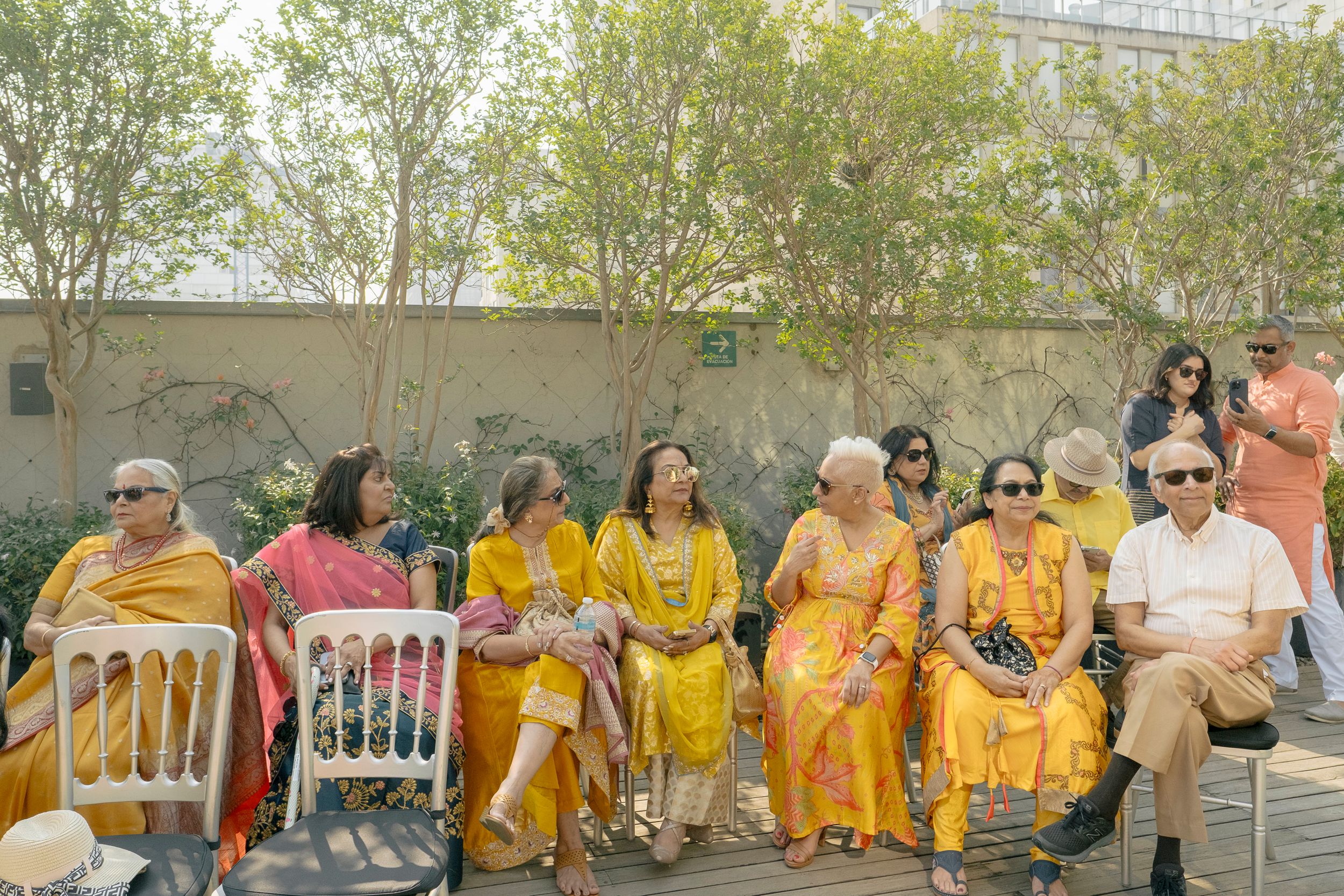Crowd gathered at an outdoor Indian cultural celebration with floral decorations and yellow attire under a hazy sky.