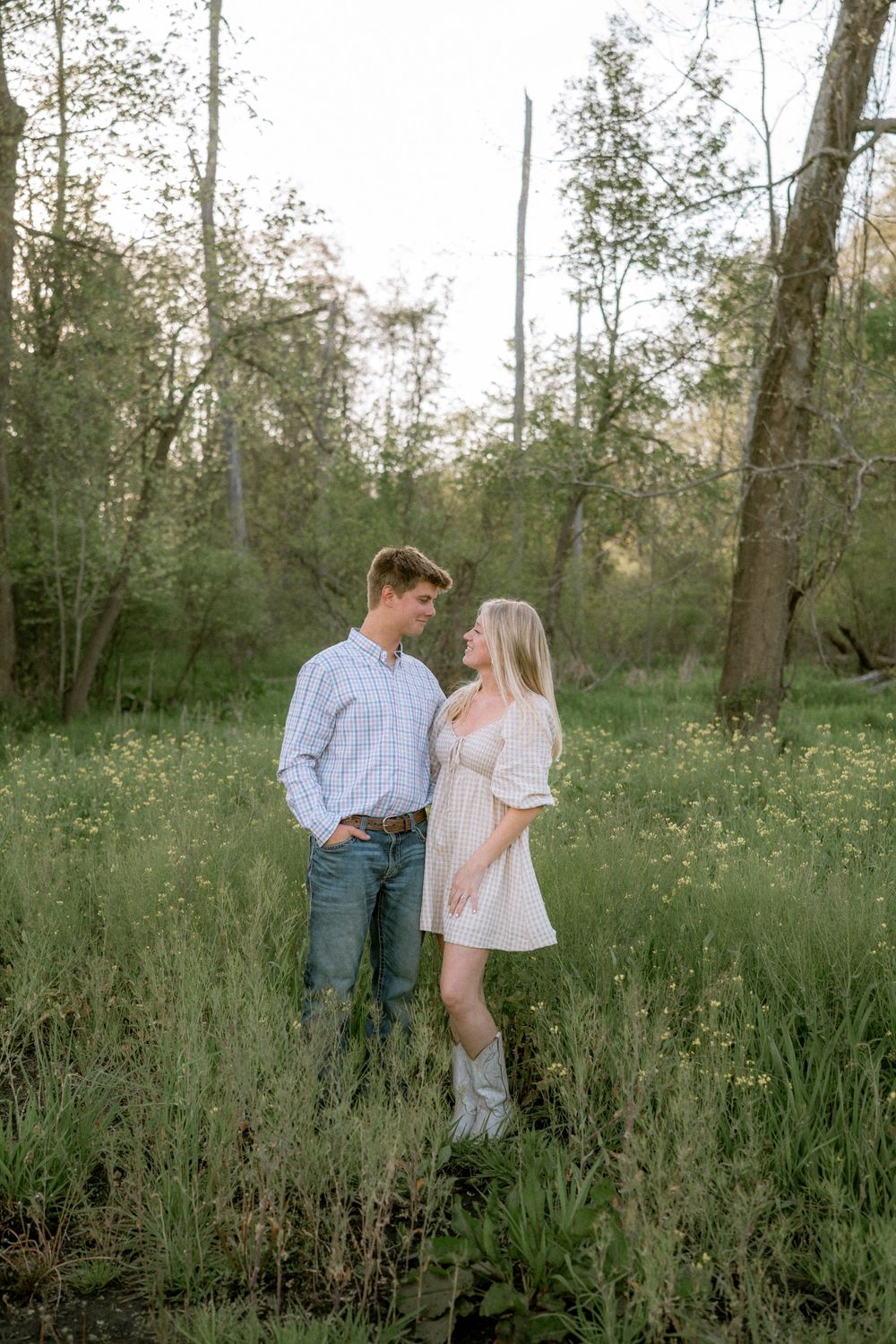 A couple stands facing each other in a lush green meadow, the woman in a white dress and boots, the man in jeans.