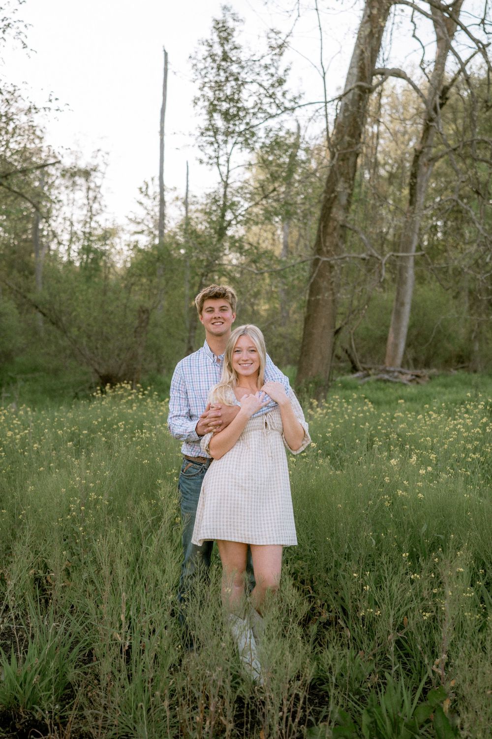 A young couple embraces in a lush green meadow with wildflowers, the man hugging the woman from behind, both smiling.