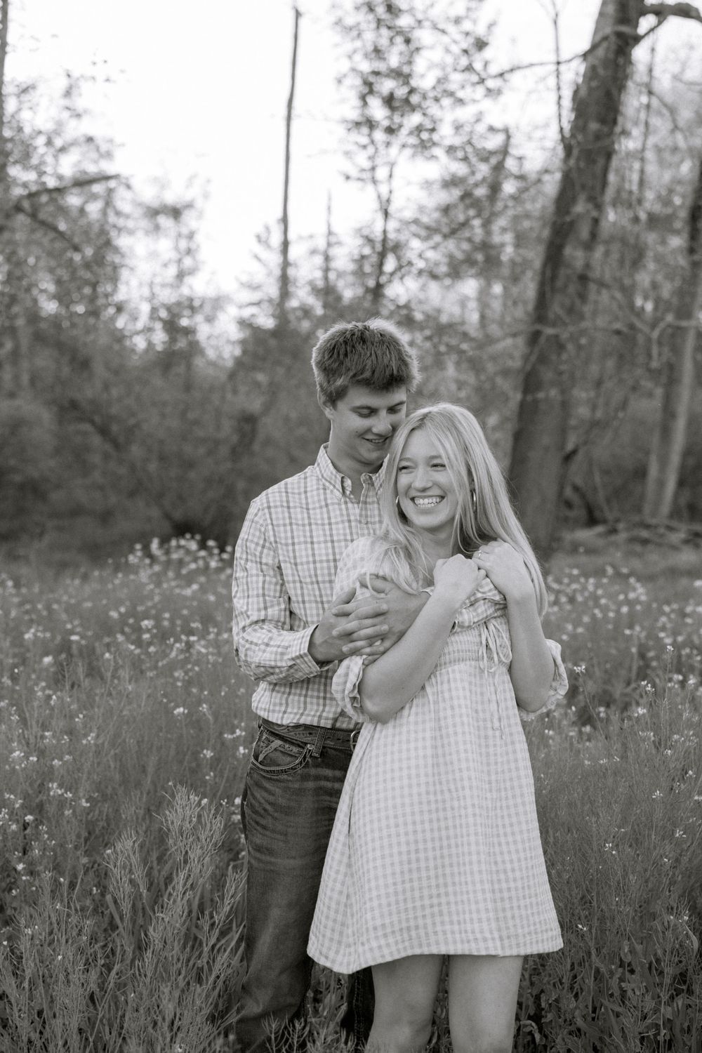 Black and white photo of a smiling couple embracing in a wildflower meadow with trees in the background.