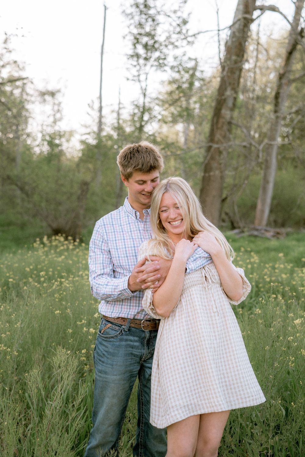 A happy couple embraces in a lush green meadow with wildflowers, the woman laughing joyfully in a plaid dress.