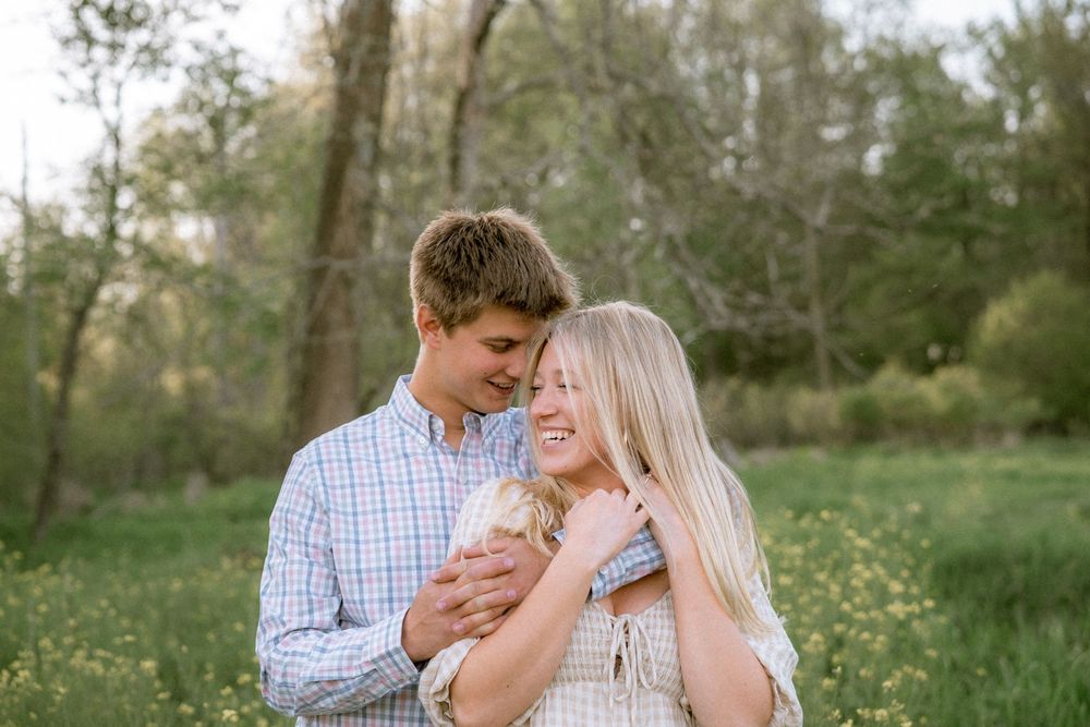 A smiling couple embraces in a sunny green meadow filled with wildflowers.