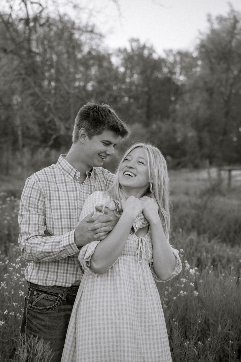 Black and white photo of a laughing couple embracing in a wildflower field at golden hour.