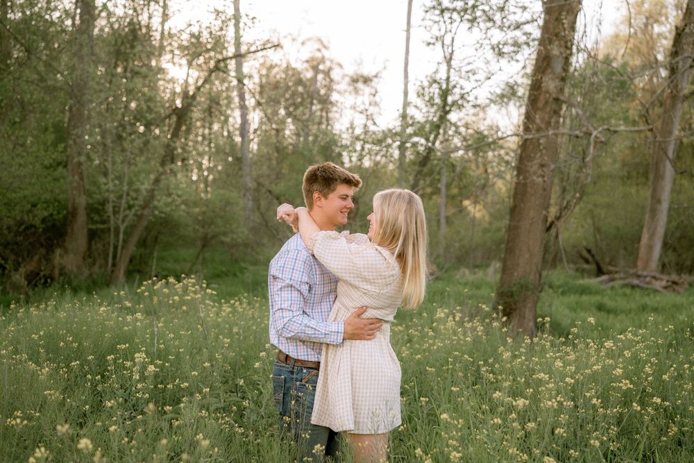Couple embracing in a lush green meadow surrounded by trees during golden hour in a romantic outdoor setting.