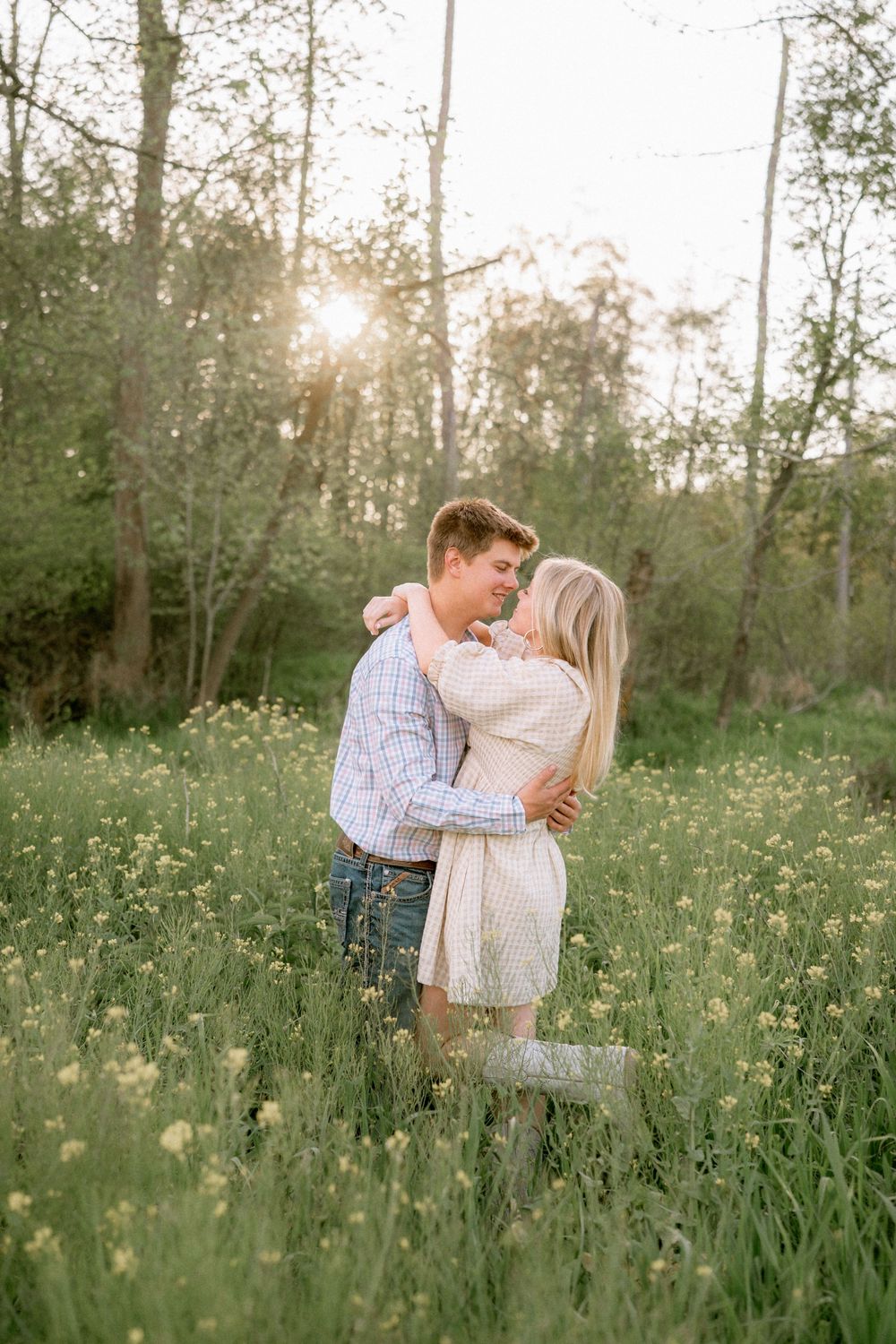 Couple embracing in a sunny wildflower meadow surrounded by trees during golden hour.