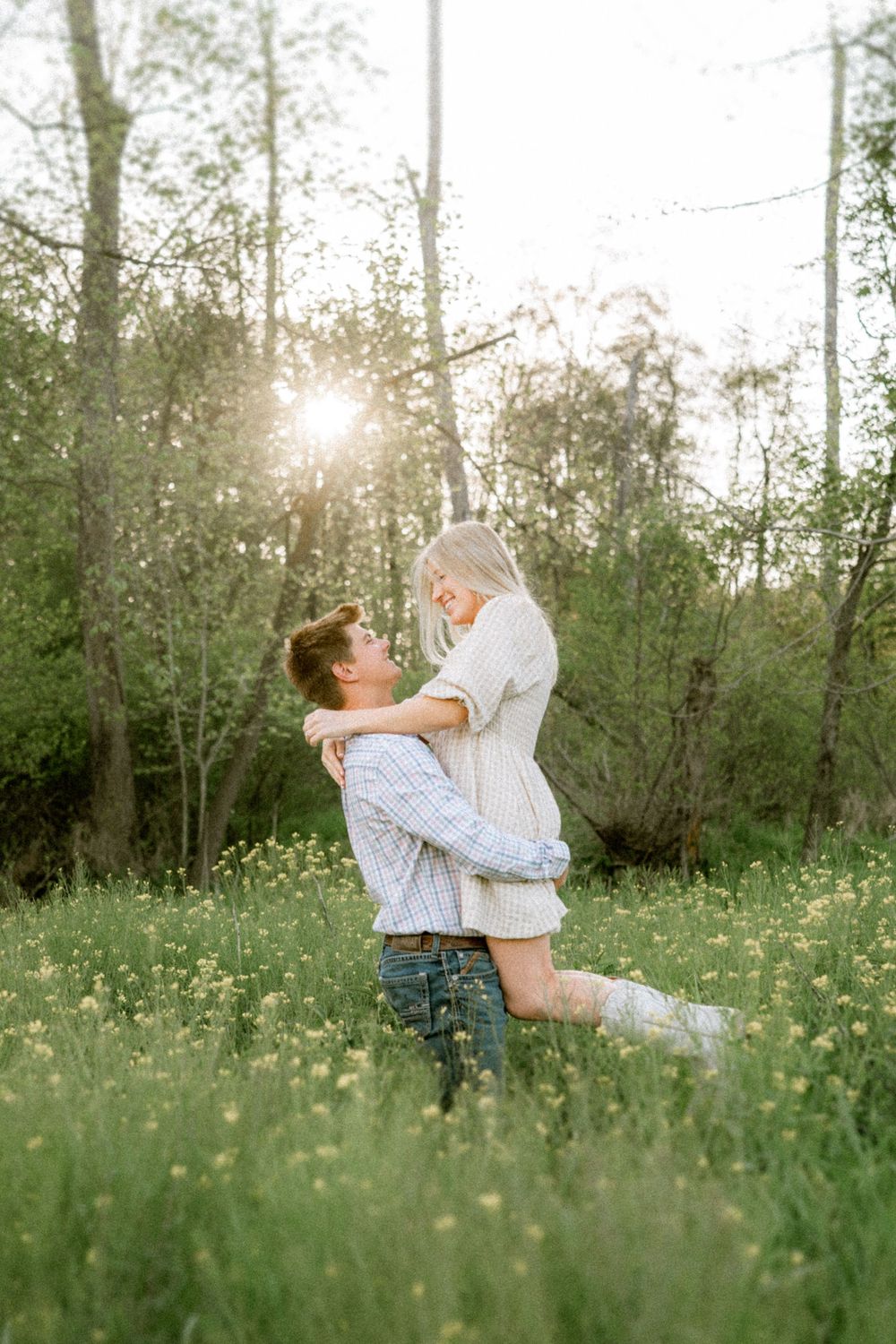 Couple embracing in sunlit meadow with wildflowers, man lifting woman joyfully among green trees.