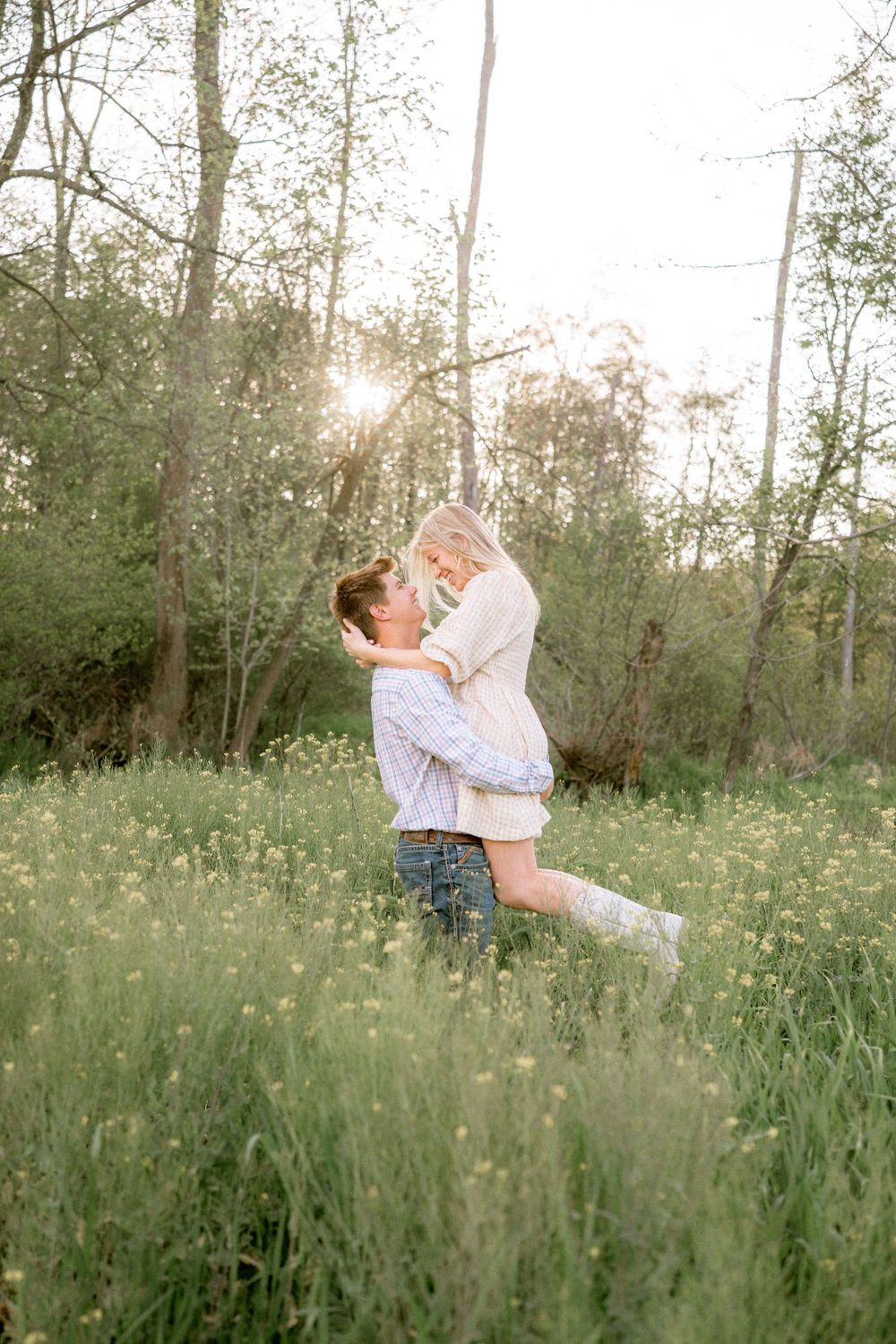 Man lifting woman in sunny meadow with wildflowers and golden backlight through trees.