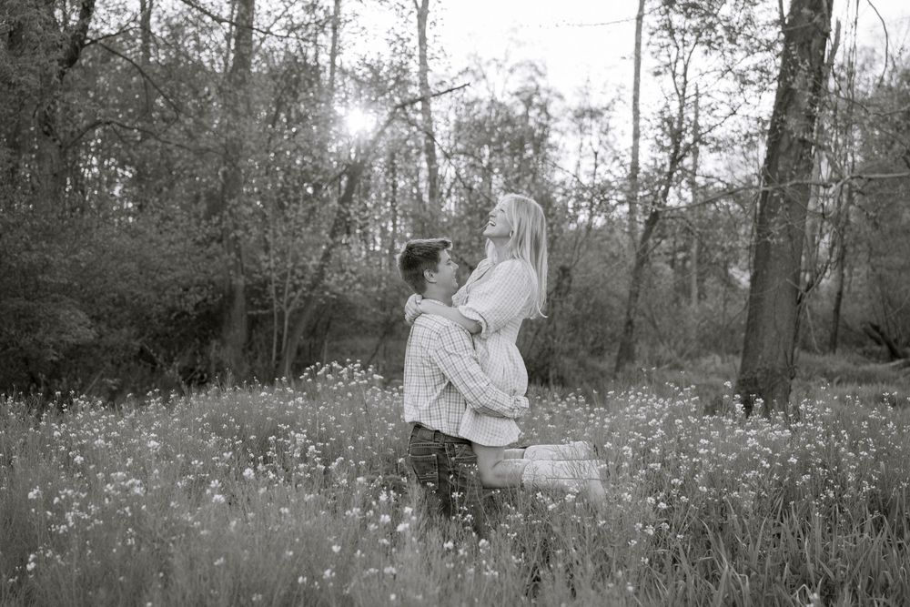 Black and white photo of a couple embracing in a wildflower meadow surrounded by tall trees.