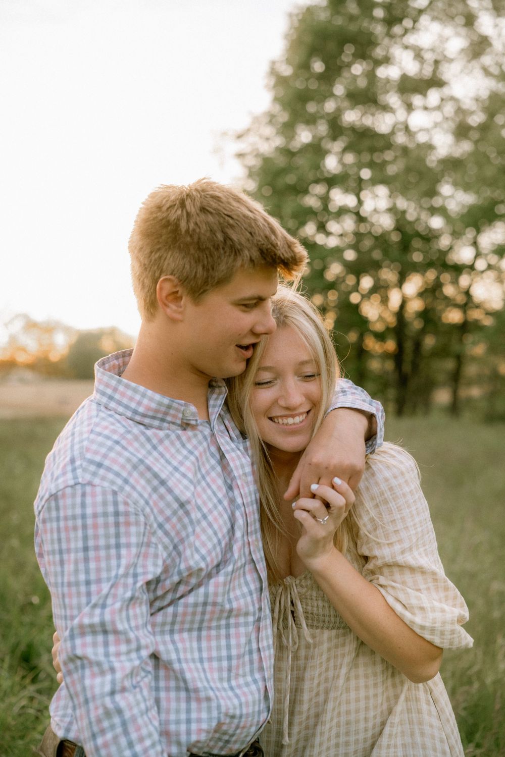 A young couple embraces and laughs together in a golden sunlit field surrounded by green trees.