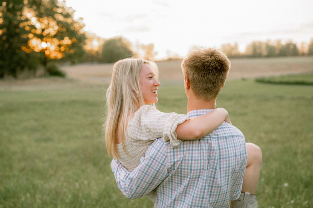 Couple embracing at sunset in a green field, woman smiling over man's shoulder in golden hour light.