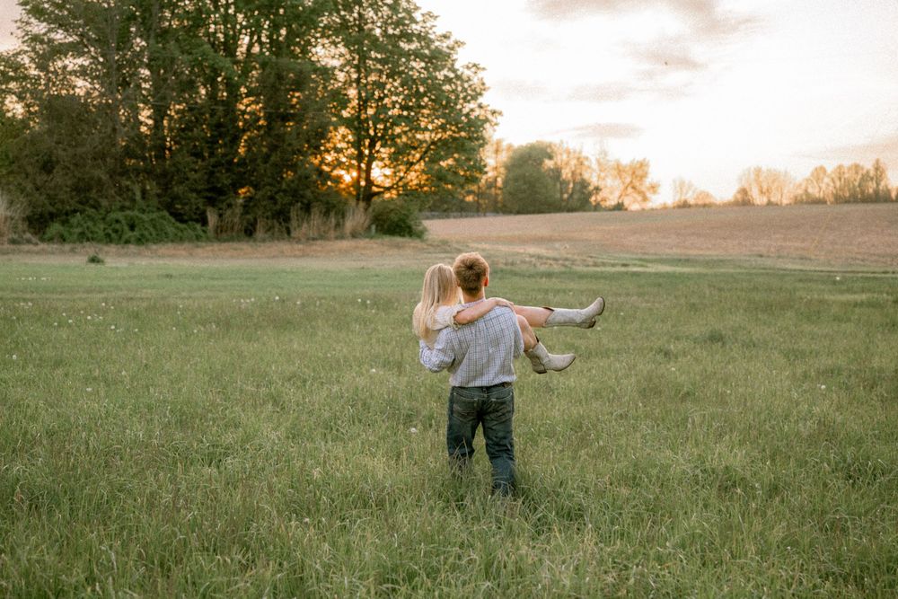A man carries a woman through a lush green field at sunset with trees in the background.