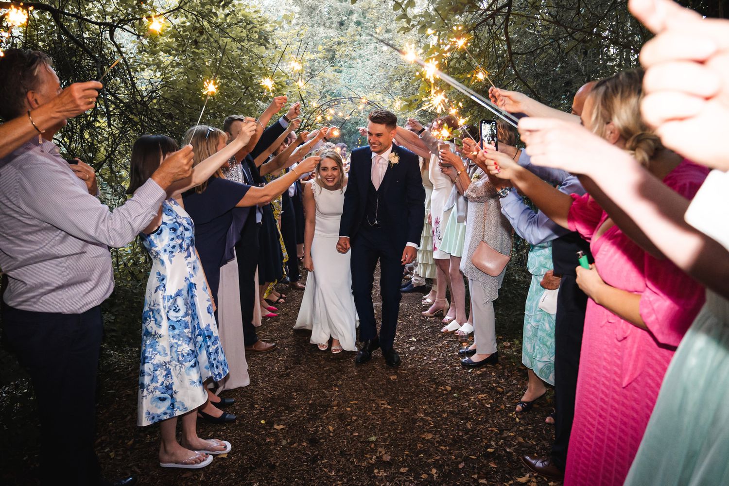 Bride and groom walk through sparkler tunnel surrounded by guests at outdoor wedding celebration.