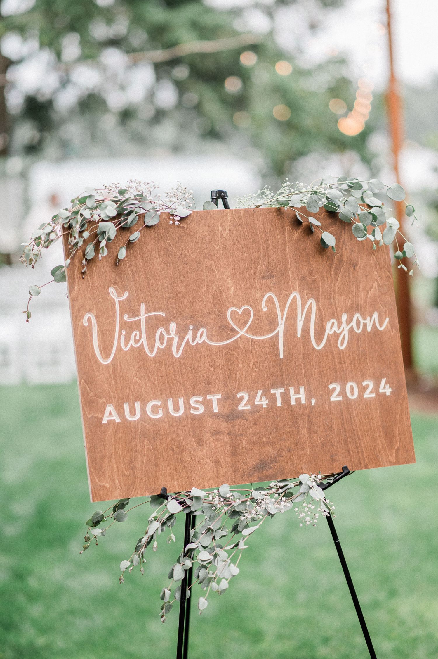 Wooden wedding welcome sign for Victoria and Mason, August 24th, 2024, decorated with eucalyptus greenery on an easel.