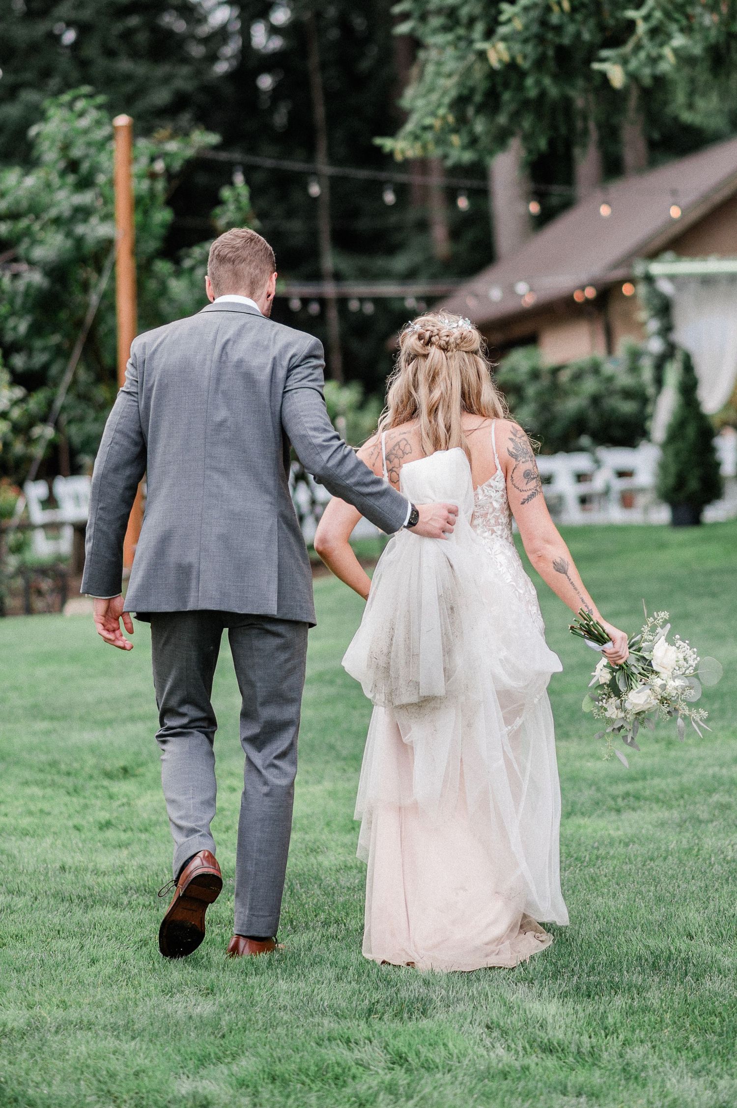 A bride and groom walk while he holds her dress across a lush green lawn at a backyard wedding surrounded by trees.