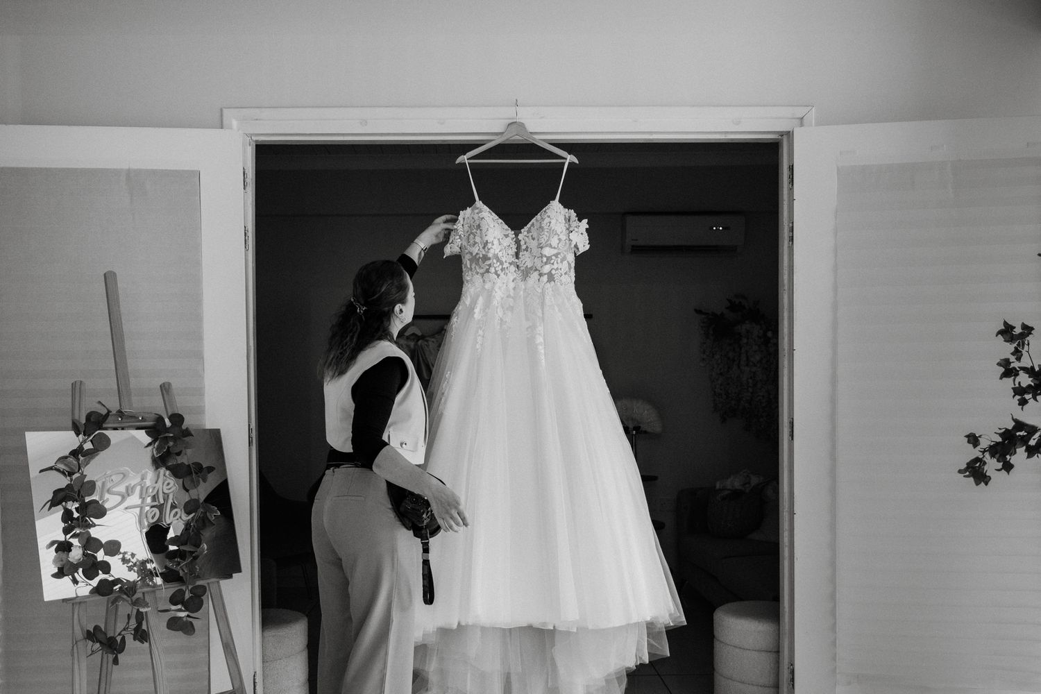 Black and white photo of a bride admiring her wedding dress hanging in a doorway before the ceremony.