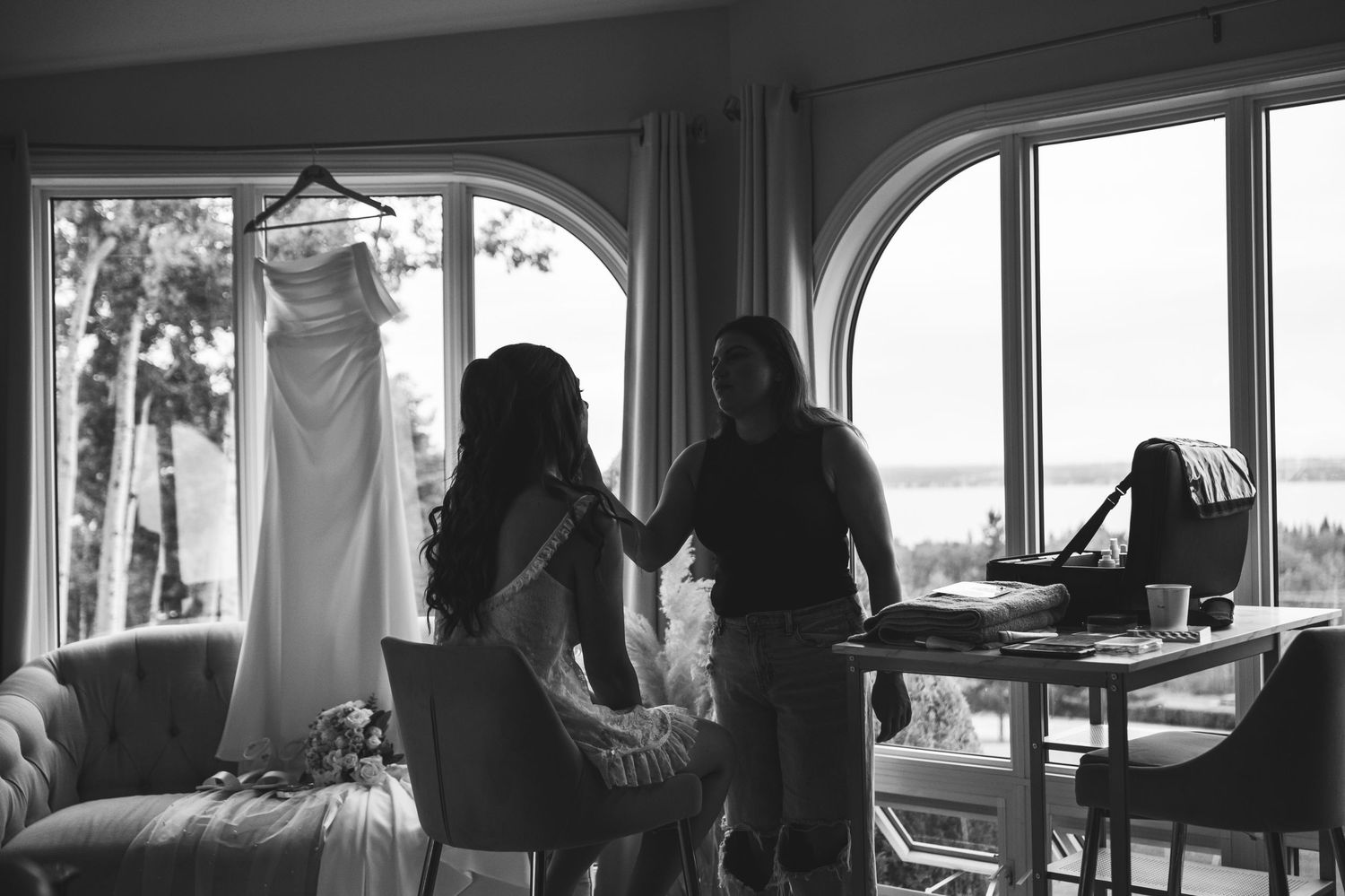 Black and white photo of bride and stylist reviewing wedding dress hanging by arched windows with scenic water view.
