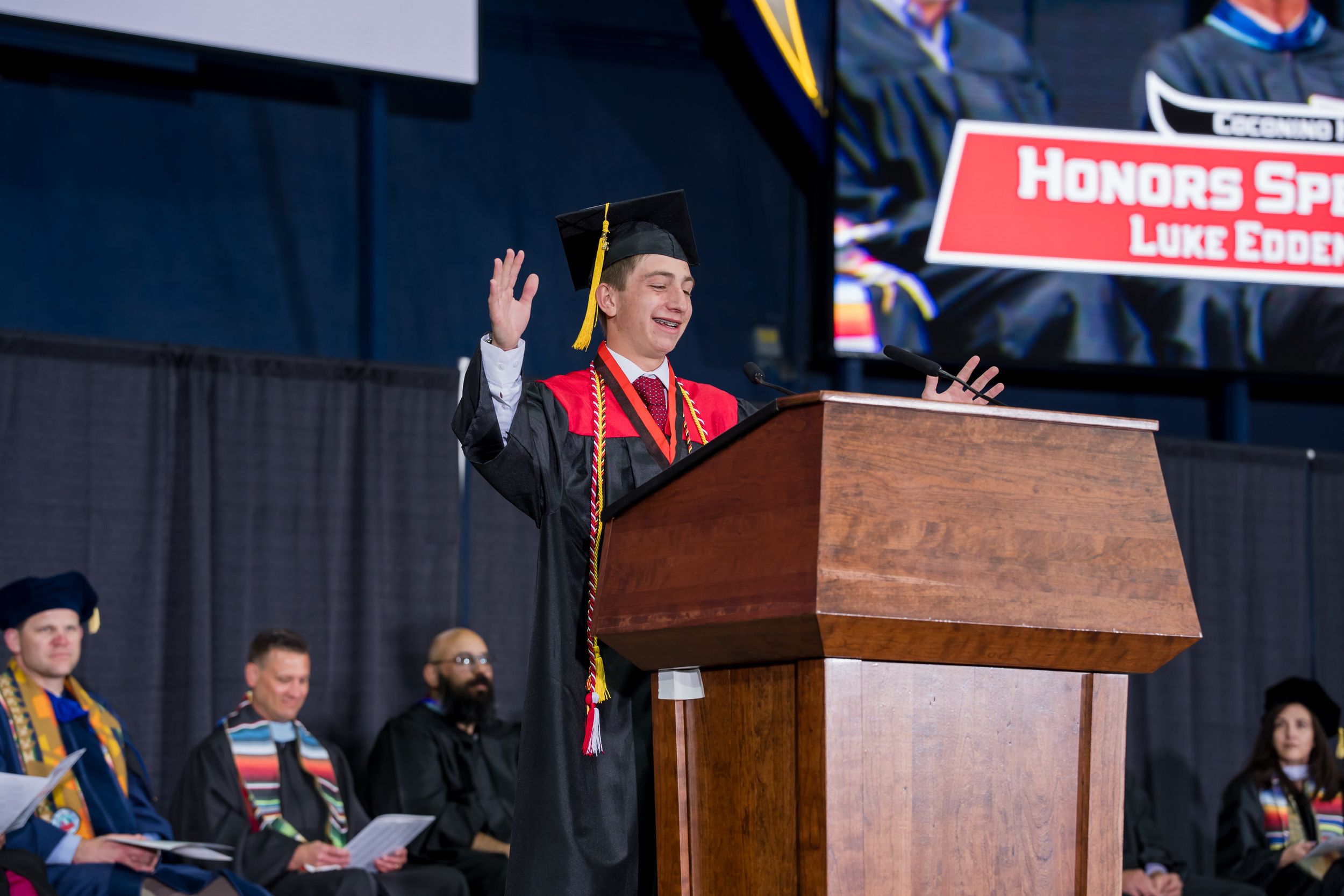 Graduate Luke Eddelman delivers honors speech at podium during university commencement ceremony in cap and gown.