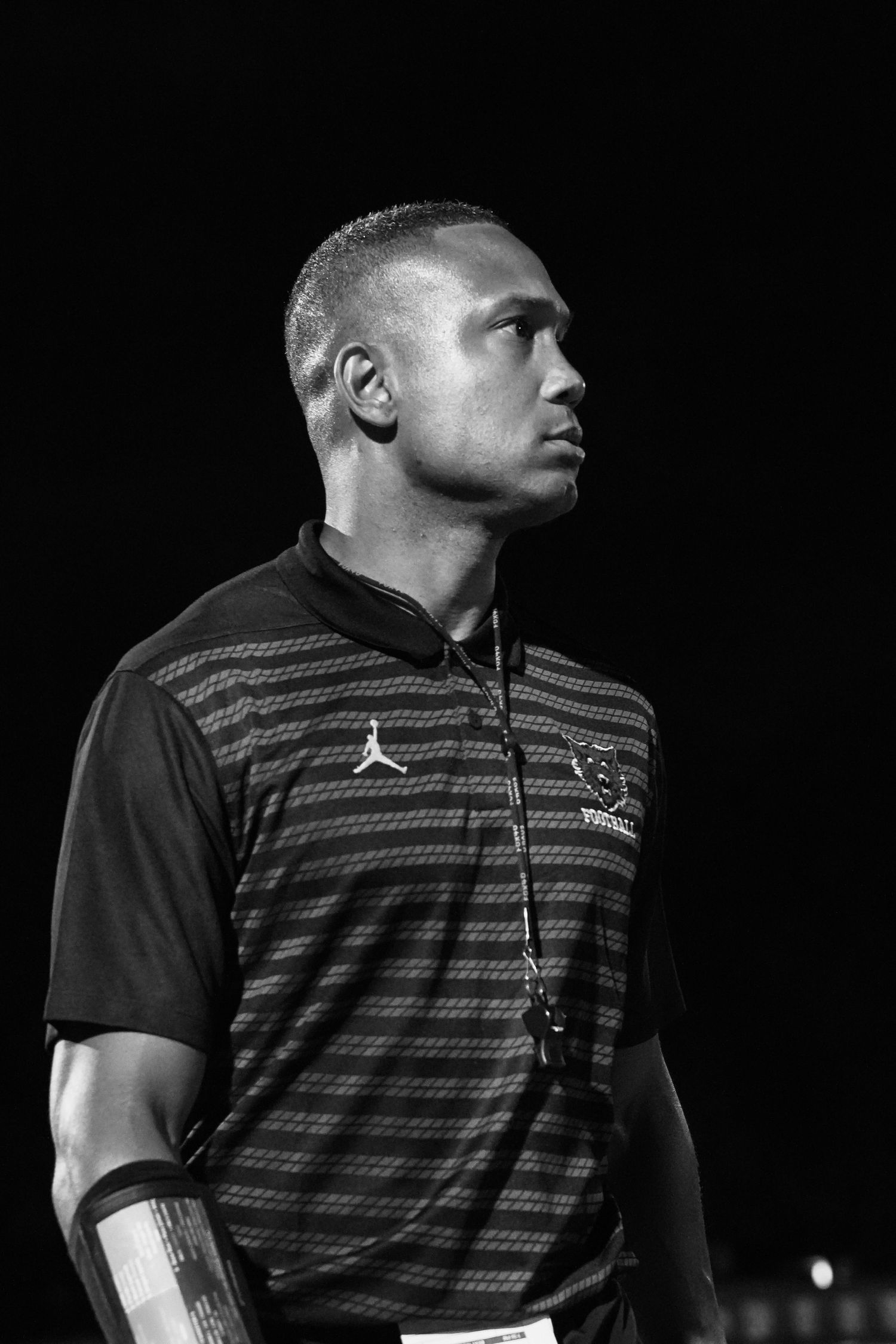 Black and white portrait of a focused man wearing a Jordan Brand polo shirt with a lanyard at a sporting event.
