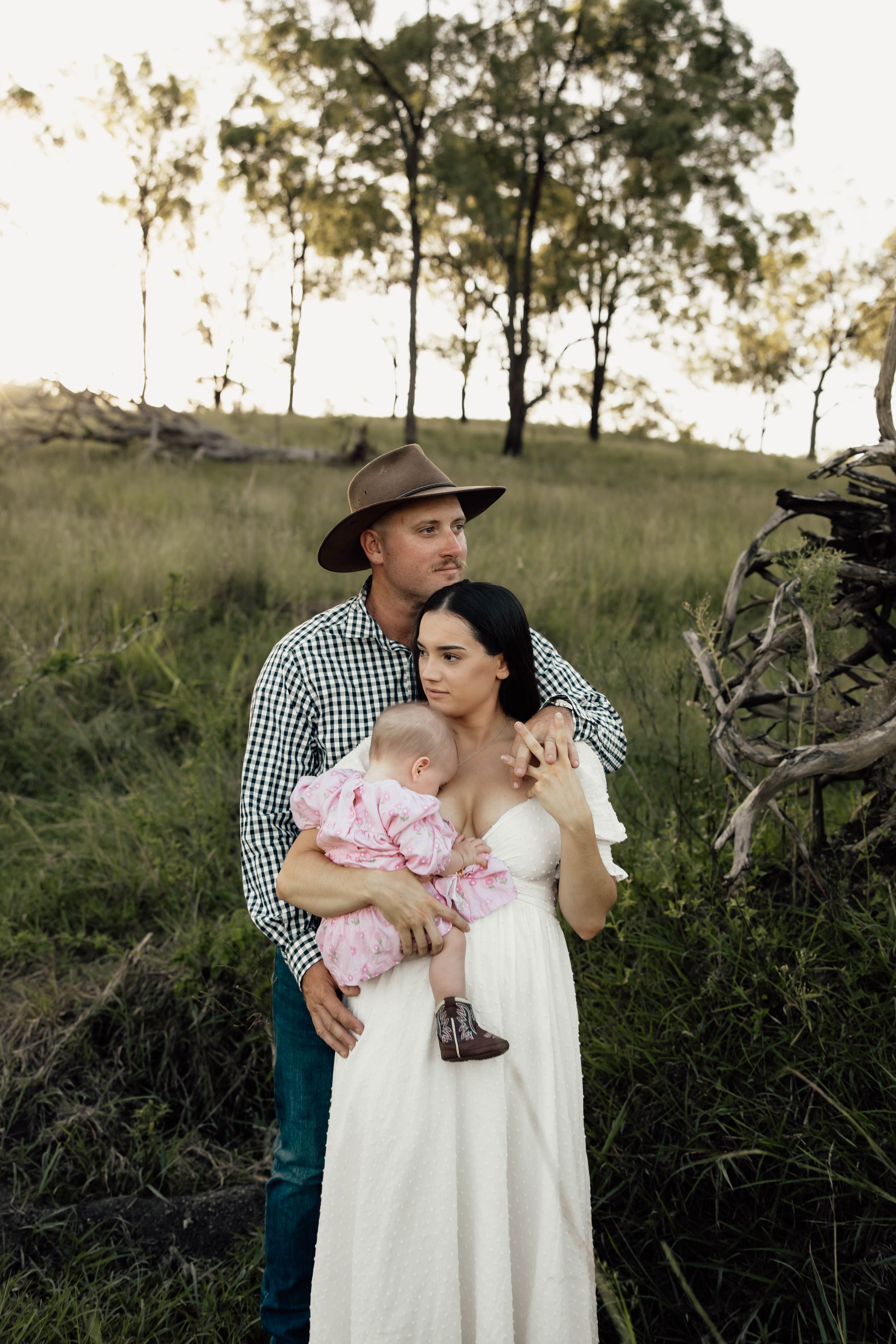 A couple in a green field holding their baby, man in cowboy hat and plaid shirt, woman in white dress.