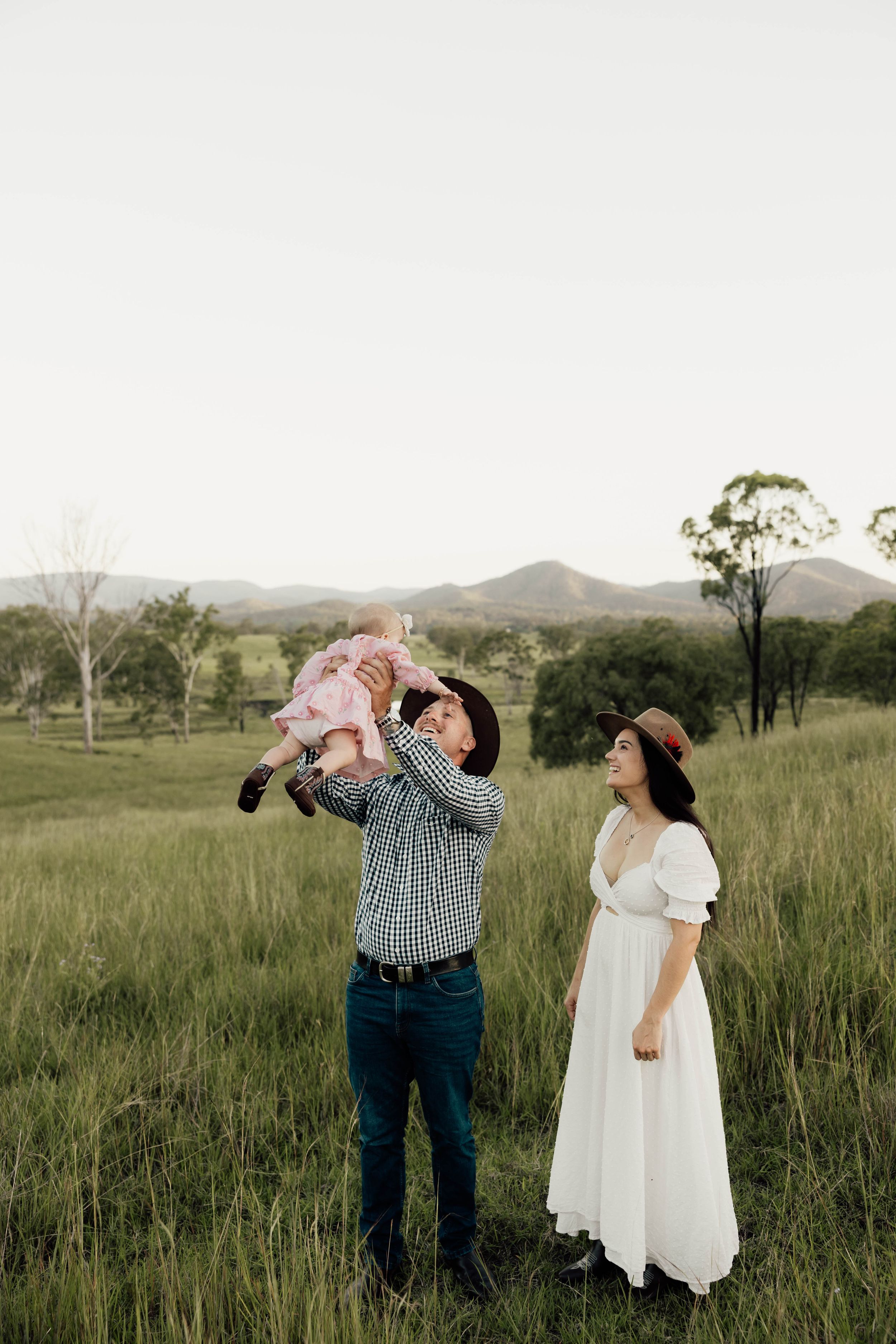 A family in a green field, dad lifting baby girl up high while mom in white dress and hat watches, mountains behind.